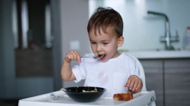 Beyaz tişörtlü şirin bebek mutfakta yüksek sandalyede oturuyor. Güzel çocuk, öğle yemeğinde çorba içiyor..