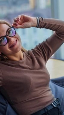 Gülümseyen mutlu bayan gözlüklerine dokunuyor. Fasulye sırığında oturmuş işten dinlenen rahat bir kadın. Arkaplan bulanık. Dikey video