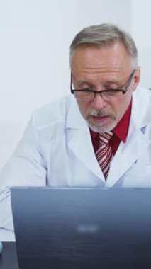 Profesyonel doktor uzak bir hastayla konuşur. Klinikte dizüstü bilgisayara bakarken görüntülü konuşma yapan beyaz üniformalı bir erkek doktor. Dikey video