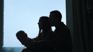 Kadın ve erkek pencere eşiğinde oturup bebek sallıyorlar. Ebeveynler ebeveynliklerinin tadını çıkarıyorlar.