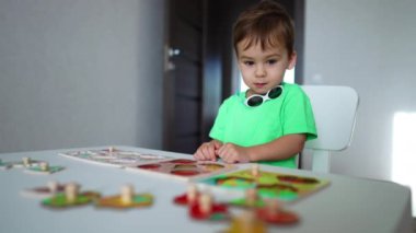 Yeşil tişörtlü sevimli çocuk evde masada oturmuş oyuncaklarla oynuyor. Gülümseyen çocuk bulmacaların parçalarını topluyor.