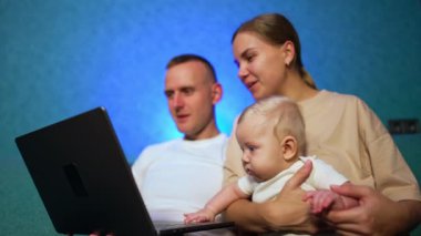 Sarışın, güzel bir bebeği olan mutlu beyaz anne ve babalar kanepeye otururlar. Gülümseyen aile dizüstü bilgisayara bakar. Düşük açı görünümü.