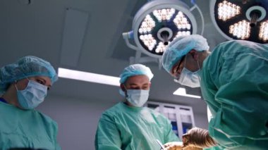 Tıbbi ekip operasyon masasında. İki erkek cerrah ve kadın hemşire yaya olarak ortopedik ameliyat yapıyorlar. Düşük açı görünümü.