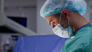 Maskeli ve şapkalı orta yaşlı erkek doktor. Ameliyat masasına bakan bir cerrahın yan görünümü.