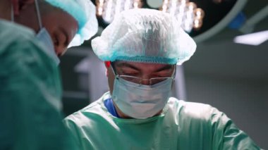 Maskeli, şapkalı ve gözlüklü yetişkin erkek doktor işine odaklanmış durumda. Ameliyathanede bir cerrahın portresi..