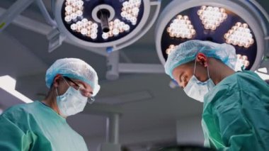 Maskeli ve şapkalı iki doktor ameliyat odasındaki yuvarlak lambaların altında duruyor. İşlemde çalışan profesyonellere düşük açı görünümü.