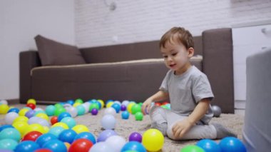 Gri tişörtlü bir çocuk odasında yerde oturuyor. Güzel çocuk, halıya saçılmış topların arasında zıplıyor..
