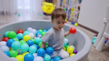 Kuru havuzda oturan sevimli siyah saçlı erkek bebek. Neşeli çocuk topları havuzdan atar..