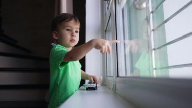 Ciddi bir beyaz çocuk pencerede durup parmağıyla cama işaret ediyor. Tatlı çocuk oyuncak arabayla oynuyor ve pencereden dışarı bakıyor..