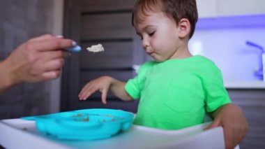 Annenin eli bütün kaşığı oğluna uzatıyor. Bebek annesinin kaşığını reddediyor ama kendi kaşığıyla yemek yiyor. Düşük açı görünümü.