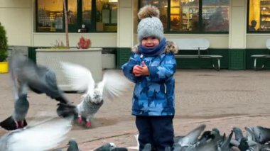 Sıcak kıyafetler içindeki sevimli erkek bebek güvercin kalabalığının yanında duruyor. Yavru kuş uçtuğunda sevinir.