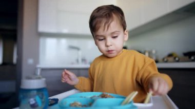 Güzel siyah saçlı erkek bebek yüksek sandalyede oturuyor. Çocuk biraz yiyecek alır, tabağa koyar ve mutlu bir şekilde alkışlar..