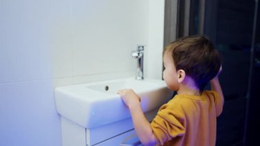 Lavabonun yanında duran siyah saçlı bebek. Bebek musluğu açar ve kapatır..