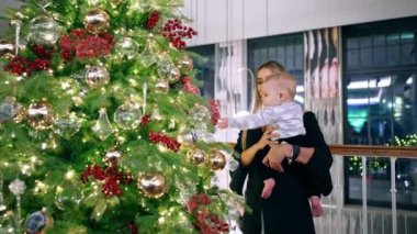 Annemin elindeki küçük bebek Noel ağacındaki parlak süslemelere ulaşmaya çalışıyor. Küçük çocuk şenlik havasına hayran kaldı..