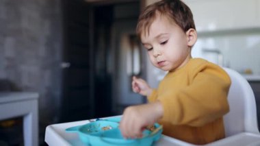 Küçük çocuk tabağına biraz yemek koyuyor. Sakin çocuk bir parça alır ve ısırır. Kapatın. Bulanık arkaplan.