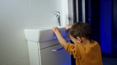Banyoda oynayan mutlu, beyaz bir bebek. Çocuk su musluğunu açar ve kapatır ve üzerinde durduğu aksesuarın yanına adım atar..