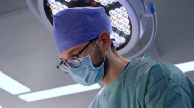 Maske takan ve gözlük takan genç bir sakallı cerrahın özenli yüzü. Düşük açı. Tavandaki doktorun arkasındaki yuvarlak lamba..