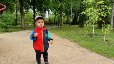 Şapkalı, spor ceketli ve pantolonlu beyaz çocuk dışarıda duruyor. Çocuk ileriye bakıyor ve işaret ediyor. Arkaplanda yeşil doğa.