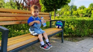 Parktaki bankta oturan küçük sevimli erkek bebek. Konuşkan çocuk şişeden su içiyor..