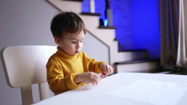 Tatlı beyaz çocuk elinde kalemle masa başında oturuyor. Küçük oğlan kalemini kağıdın yanına koyuyor..