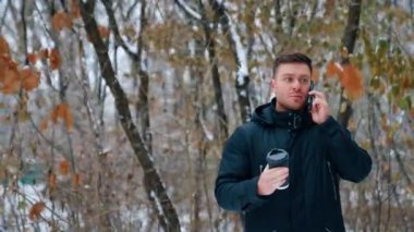 Siyah ceket giyen yakışıklı, beyaz bir erkek telefonda konuşuyor. Elinde kahve fincanı olan bir adam kışın dışarıda telefonla konuşuyor..