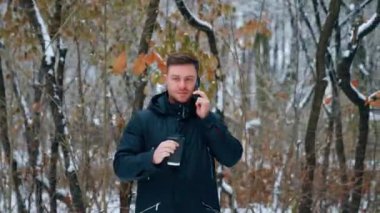 Koyu renk saçlı beyaz erkek kahve içer ve telefonda konuşur. Kış parkında yürüyen bir adam..