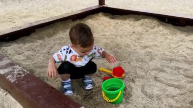 İki yaşındaki sevimli çocuk bir kova suyla oynuyor. Sevimli çocuk kum havuzunda otururken el çırpıyor ve ıslak eller ovuyor..