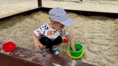 Küçük çocuk kum havuzunda oturup oyuncaklarıyla oynuyor. Gülümseyen çocuk oyuncak kovasından su sıçratıyor..
