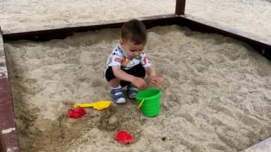 Beyaz, şirin bir çocuk kum havuzunda çömelmiş oturuyor. Çocuk bir avuç kum alıyor, ağzına çekiyor ve kovaya koyuyor..
