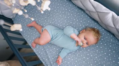 Mavi elbiseli sevimli erkek bebek beşikte yatıyor. Ağzında emzik olan tatlı çocuk bebek karyolasına bakıyor. Üst görünüm.