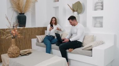 İnsanlar kanepede aletlerle otururlar. Adam dizüstü bilgisayarda çalışıyor ve kadın ona akıllı telefonunu gösteriyor..