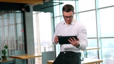 Beyaz tişört giyen Kafkas işadamı bir I-pad tutuyor ve kahve içiyor. İşi birleştiren ve ofiste ayrılan adam..