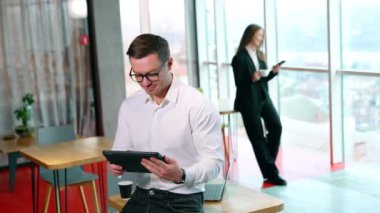 Gözlüklü adam I-pad 'ine odaklanmış görünüyor. Adam masadan bir kağıt bardak alır ve kahve içer. Siyah takım elbiseli kadın bulanık arka planda duruyor ve çay içiyor..