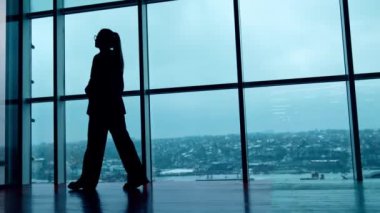 Gözlüklü ve takım elbiseli bir kadının silüeti panoramik pencereler boyunca yürüyor. Arka planda şehir panoraması. Düşük açı görünümü.
