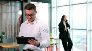 Gözlüklü bir adam kağıt bardak ve I-pad tutuyor. İşadamı aletlere odaklanmış durumda. Kadın arka planda telefonla konuşuyor..