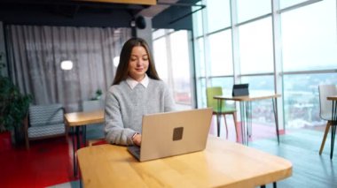 Masada uzun kahverengi saçlı, odaklanmış beyaz kadın oturuyor. Ofis çalışanı bilgisayarına yazıp gülümsüyor..