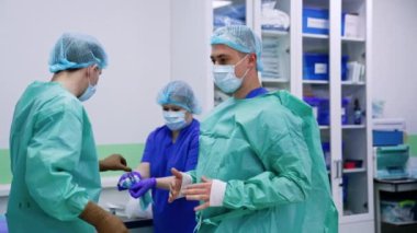 Orta yaşlı beyaz erkek doktor ameliyat için giyinmiş. Kadın hemşire gelir ve bornozu arkaya bağlar..