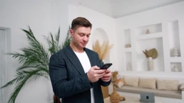 Koyu saçlı, beyaz ceketli erkek akıllı telefonuna bakıyor. Uzaktan çalışan kendinden emin bir girişimcinin portresi.