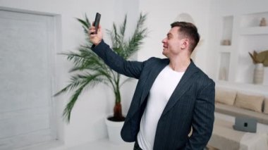 Büyük, rahat odada selfie çeken bir adam. Ceket giyen erkek telefonu başının üstünde tutuyor, gülümsüyor..