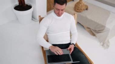 Beyaz gömlekli yakışıklı erkek dizüstü bilgisayarı açar ve çalışmaya başlar. Girişimcilerin evden bağımsız çalışmalarına ilişkin en iyi bakış açısı.