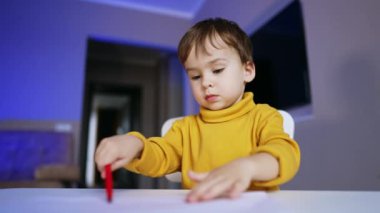 Sevimli siyah saçlı bebek evde huzur içinde resim çiziyor. Sarı kazaklı sevimli çocuk kırmızı kalem kullanıyor. Düşük açı görünümü.