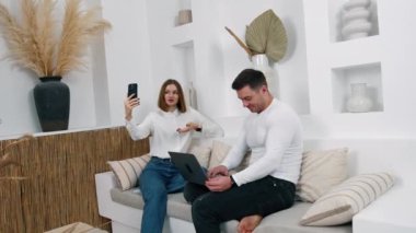 Elinde telefon olan bir kadın görüntülü konuşma yaparken selfie çekiyor. Yanında oturan adam dizüstü bilgisayarına odaklanmış..