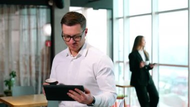 Kahve molasında çalışmaya devam etmek. Adam elinde kağıt bardakla I-pad 'e odaklanmış durumda. Bulanık arkaplanda elinde fincanla duran kadın telefonda konuşuyor..