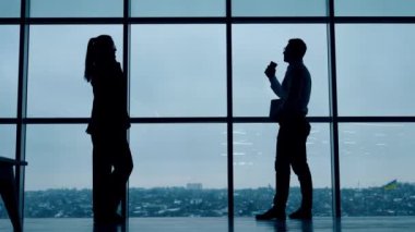 Erkek silueti pencerede duruyor ve kahve içiyor. Kadın silueti telefonda konuşurken yürür. Ofis çalışanları iş başında.