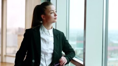 Ofisin penceresinde duran siyah ceketli beyaz kadın. İş kadını kameraya gülümseyerek gözlüğünü takar..