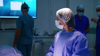 Üniforma giyen, maske takan ve ameliyat masasındaki ışıkta duran kadın doktor. Bir grup hastane personeli ameliyathanede çalışıyor..