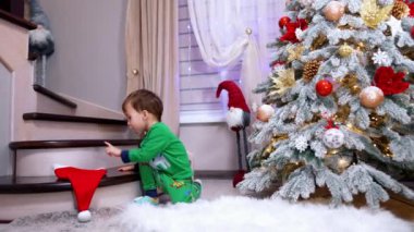 Merdivenlerden atlayan yeşil takım elbiseli küçük bir çocuk. Kırmızı Noel Baba şapkası yanında duruyor. Güzel Noel ağacı yanında duruyor.