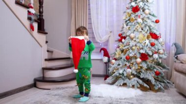 Yeşil pijama giyen çocuk güzel bir Noel ağacı olan odada duruyor. Bebek Noel Baba şapkasını takıyor.