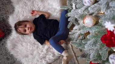 İki yaşında bir çocuk yerde yumuşak, bembeyaz bir ekose üzerinde yatıyor. Küçük çocuk Noel ağacına bakıyor. Dikey ekran.