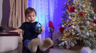 Elinde bardak olan küçük bir çocuk merdivenlerde oturuyor. Noel ağacı olan tatlı bir çocuk var odada..
